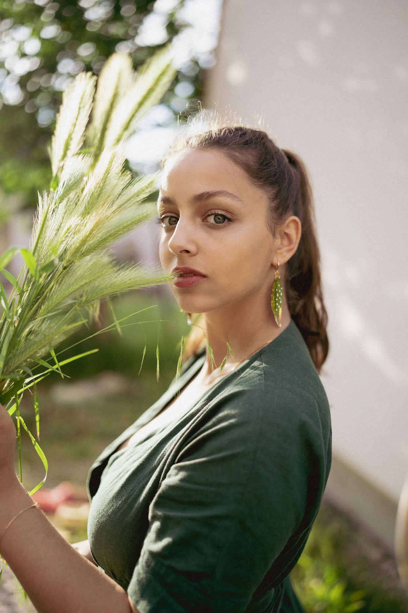 A woman with long brown hair in a ponytail, wearing a dark green top and unique jewelry—Begonia Leaves Earrings—stands outdoors in sunlight holding tall grass, looking directly at the camera. The softly blurred background is perfect for plant lovers.