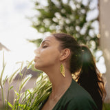 A woman with long dark hair in a ponytail stands outdoors, eyes closed, holding green plants. Sunlight softly illuminates her face as she wears unique jewelry—a large Begonia Leaves Earring. A blurred tree appears in the background, perfect for plant lovers.