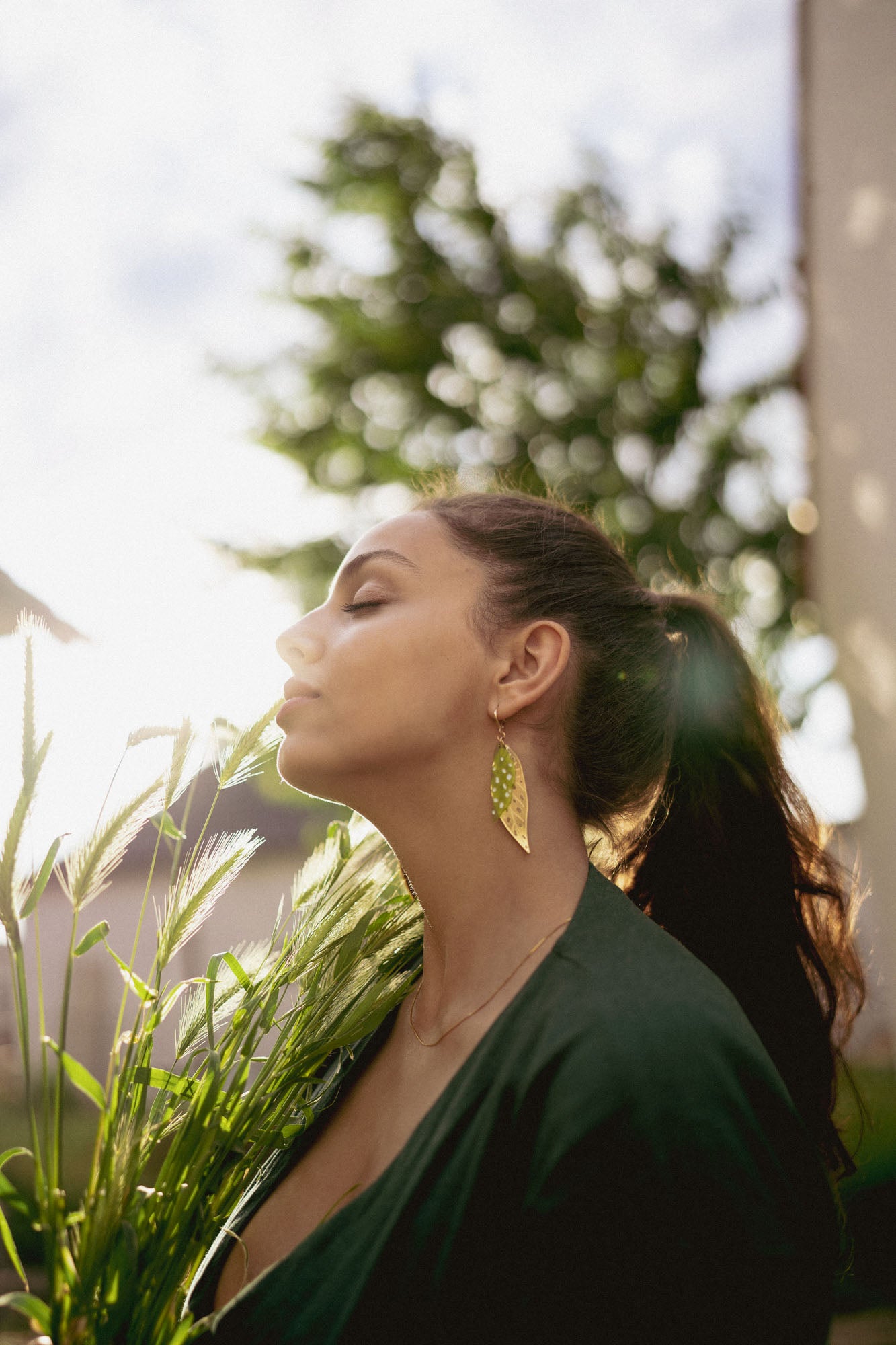 A woman with long dark hair in a ponytail stands outdoors, eyes closed, holding green plants. Sunlight softly illuminates her face as she wears unique jewelry—a large Begonia Leaves Earring. A blurred tree appears in the background, perfect for plant lovers.