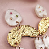 Close-up of abstract dangle earrings with textured gold shapes and marbled white pieces, decorated with small pink, green, and amber rhinestones, displayed on soft, light pink fabric.