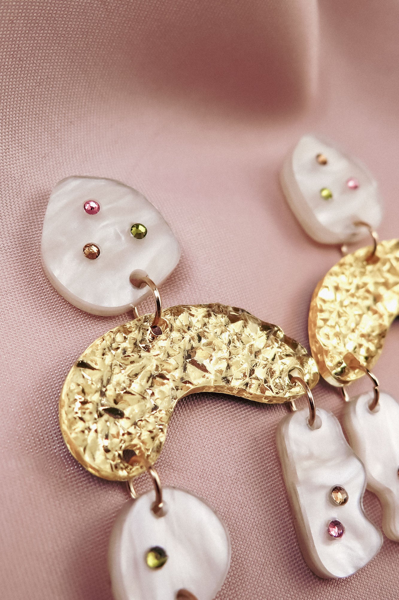 Close-up of abstract dangle earrings with textured gold shapes and marbled white pieces, decorated with small pink, green, and amber rhinestones, displayed on soft, light pink fabric.