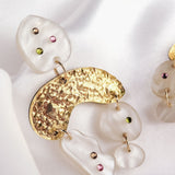 A close-up of a boho chic dangle earring featuring textured gold metal and irregularly shaped white resin pieces adorned with small pink and green rhinestones, set against a soft white satin background.