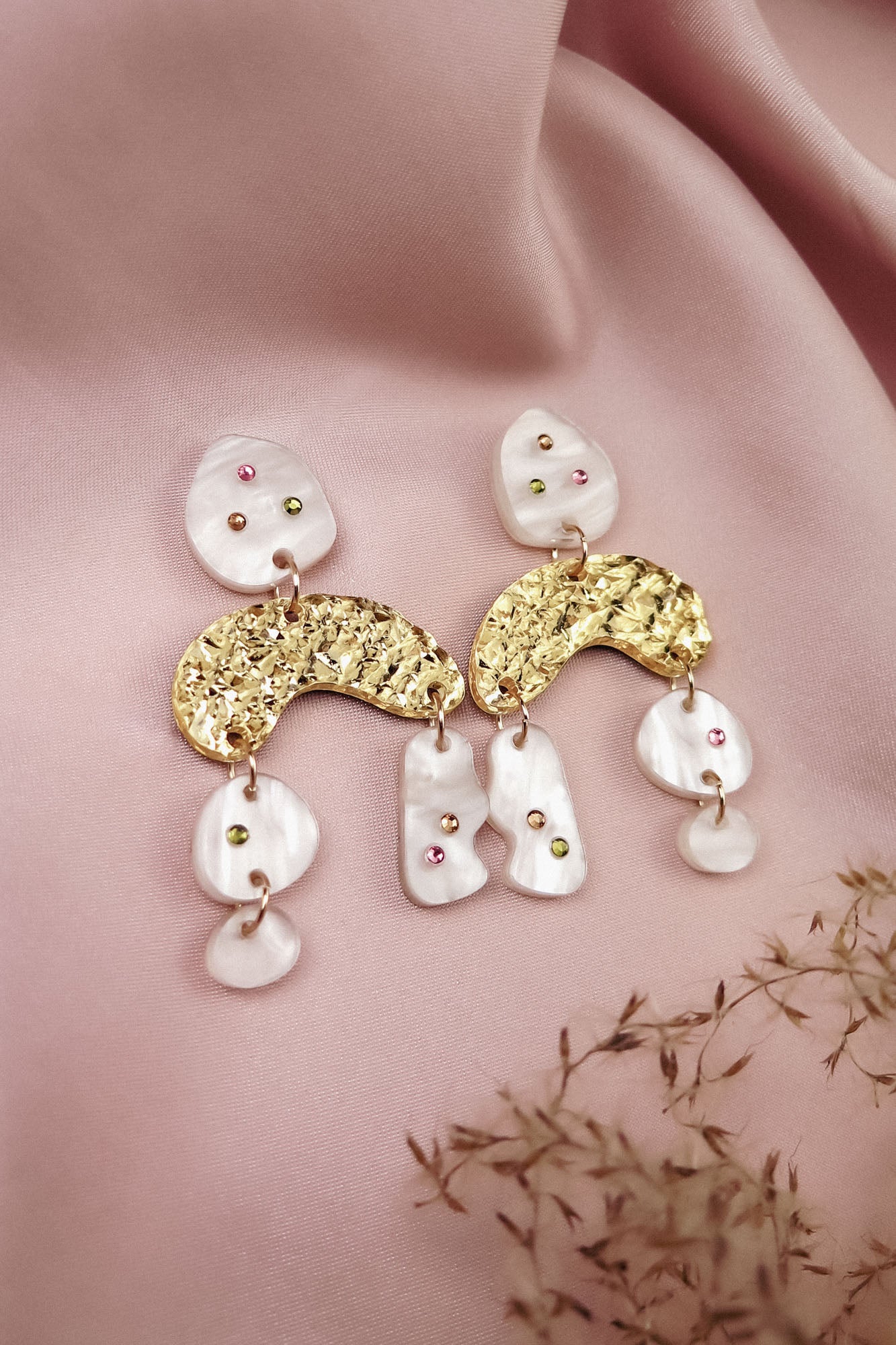 A pair of boho chic dangle earrings featuring gold textured arches and white, marble-like pieces, adorned with small colored gems, rhinestones, and displayed on soft pink fabric with dried flowers beside them.