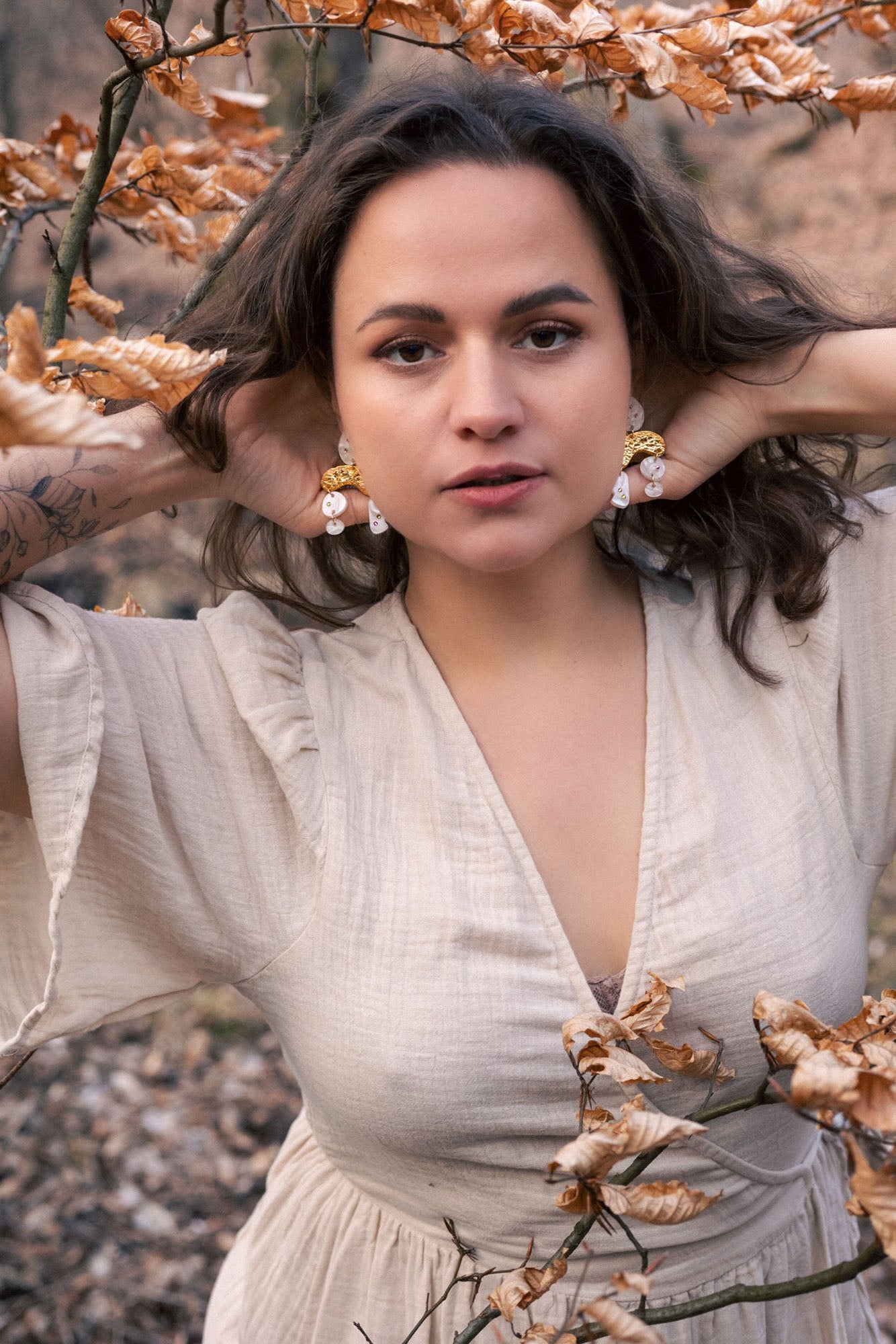 A woman with wavy brown hair poses outdoors among dry autumn leaves, wearing a light beige dress and boho chic dangle earrings adorned with rhinestones, her hands touching her hair and an intense, confident expression on her face.