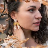A woman with wavy brown hair wearing a cream top and boho chic dangle earrings stands among branches with dry, light brown leaves, gazing thoughtfully to the right.