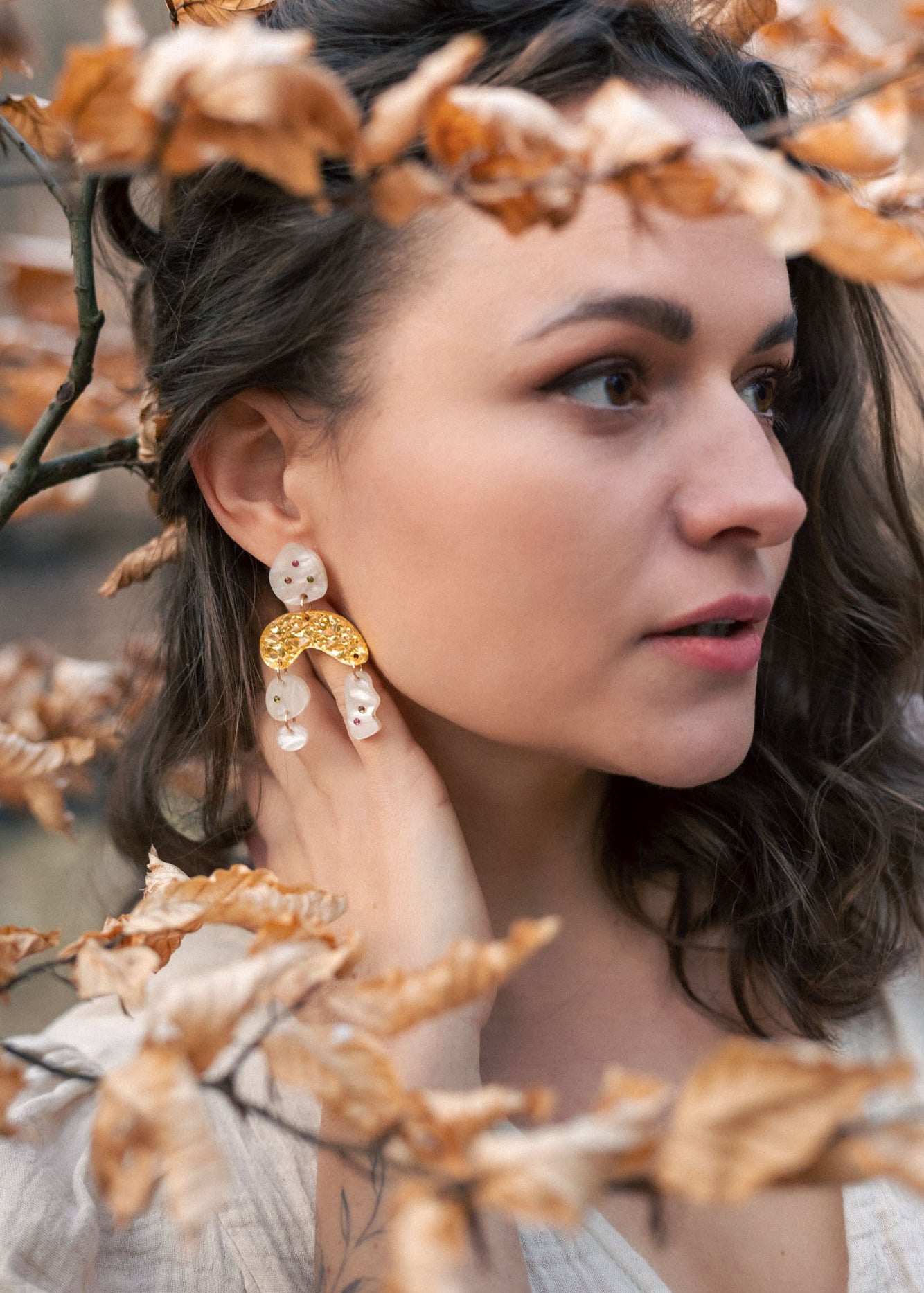 A woman with wavy brown hair wearing a cream top and boho chic dangle earrings stands among branches with dry, light brown leaves, gazing thoughtfully to the right.