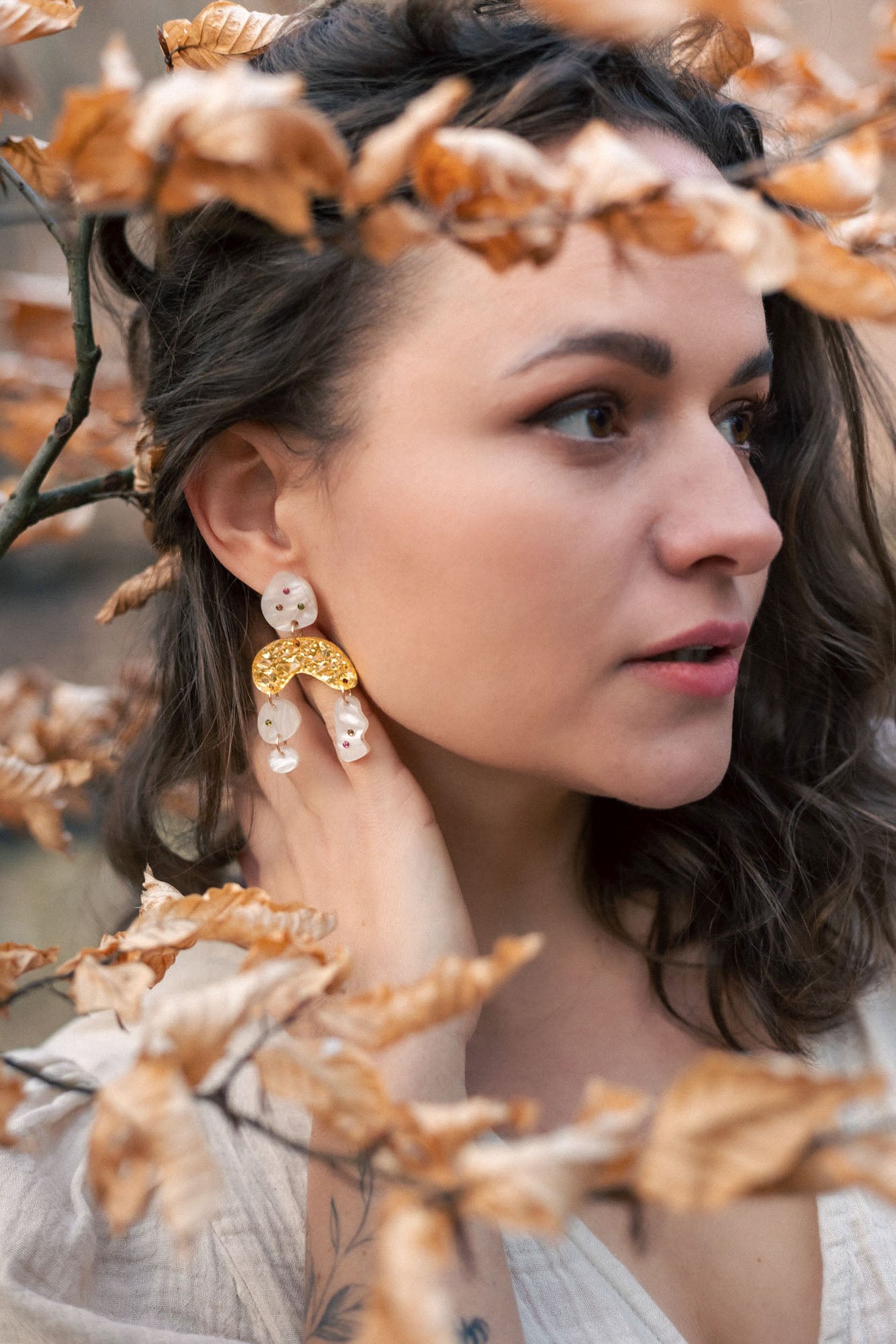 A woman with wavy brown hair wearing a cream top and boho chic dangle earrings stands among branches with dry, light brown leaves, gazing thoughtfully to the right.