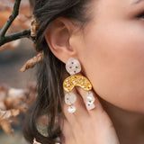 A close-up of a womans ear wearing unique boho chic dangle earrings with gold, pearl-like abstract shapes, and subtle rhinestones. Her hand touches her neck, surrounded by brown autumn leaves and branches.