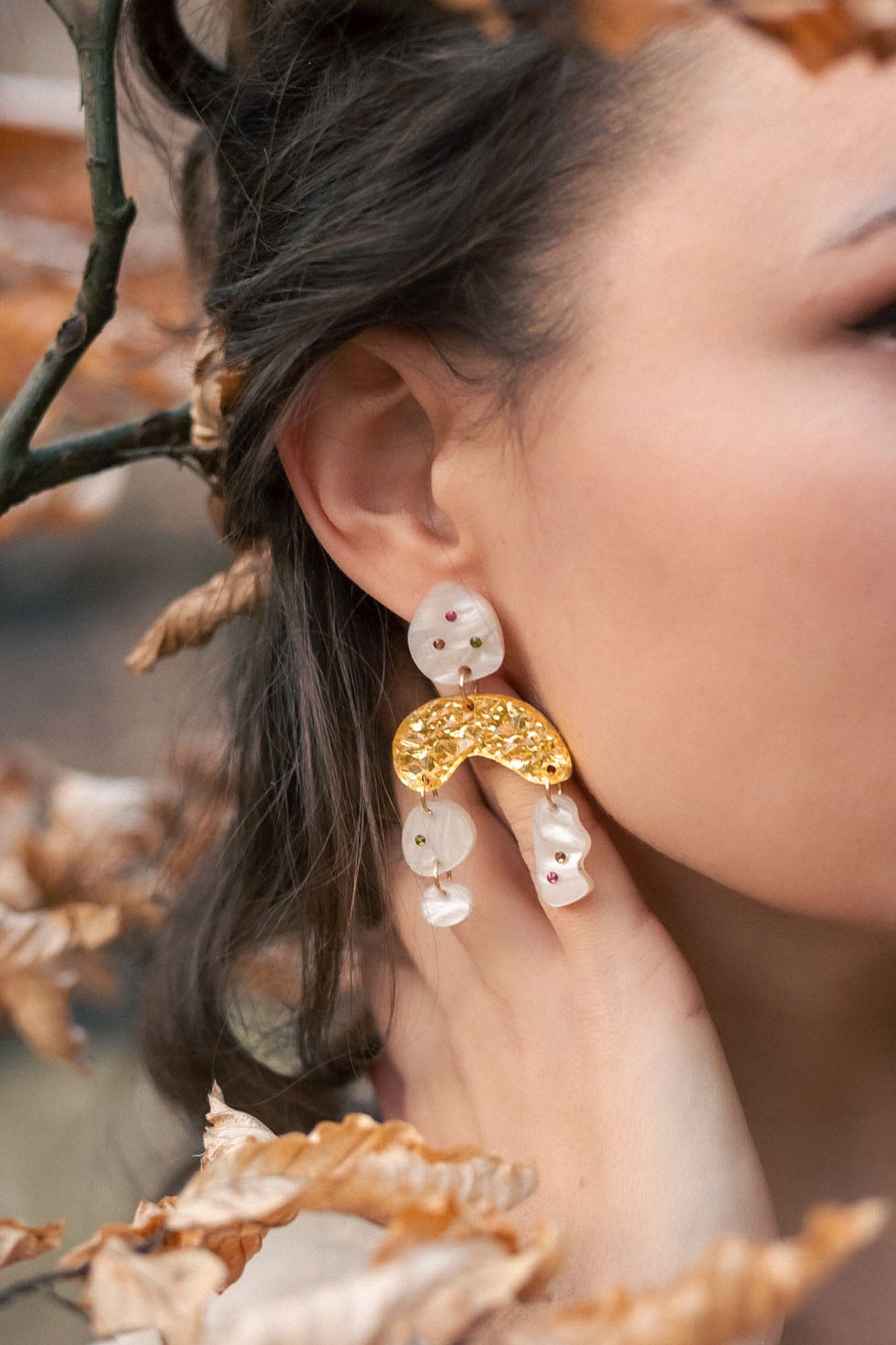 A close-up of a womans ear wearing unique boho chic dangle earrings with gold, pearl-like abstract shapes, and subtle rhinestones. Her hand touches her neck, surrounded by brown autumn leaves and branches.