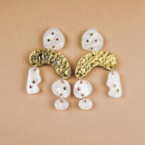 A pair of abstract dangle earrings with textured gold curved pieces, irregular white shapes, and small embedded red and green rhinestones, displayed on a beige background.