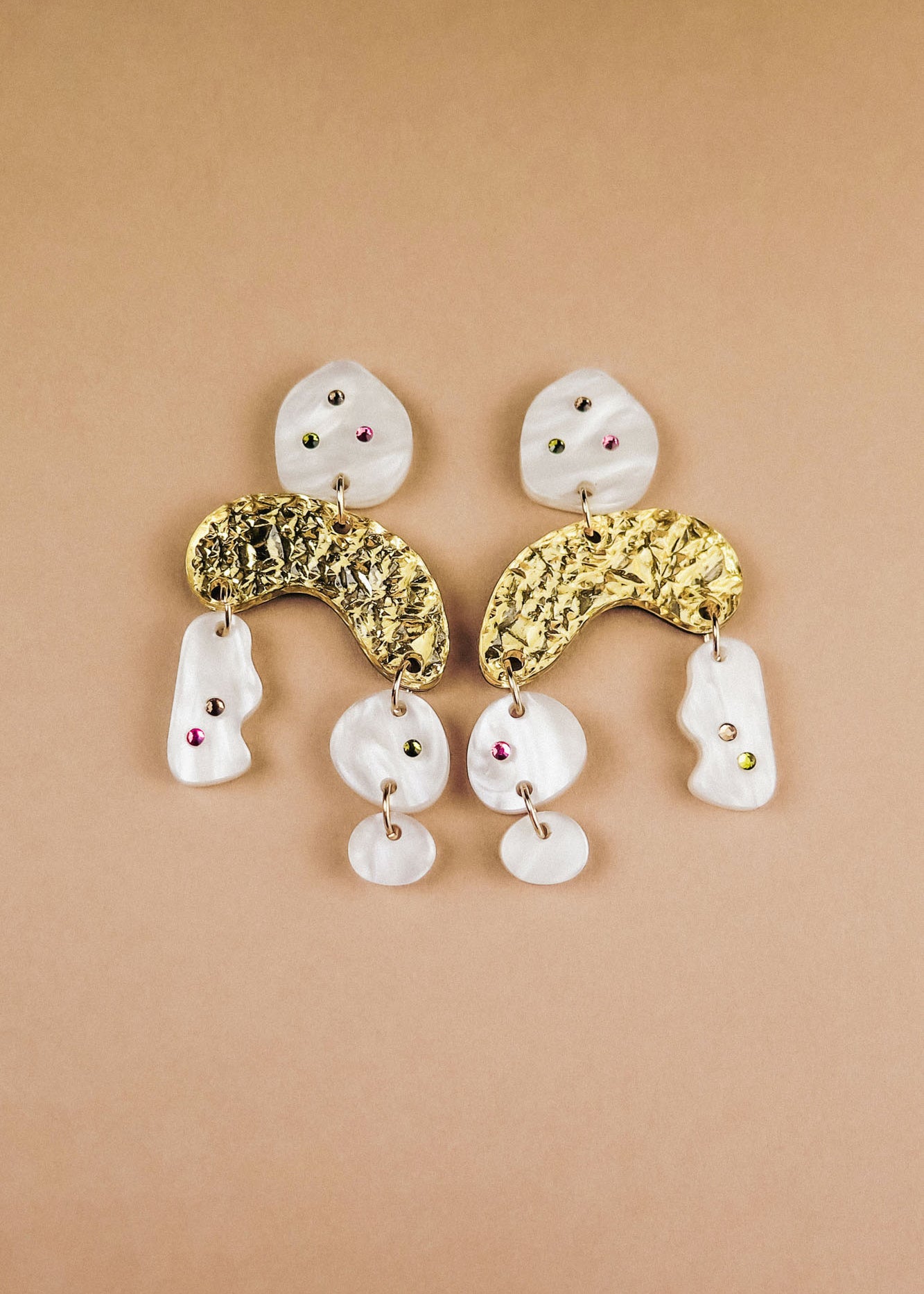 A pair of abstract dangle earrings with textured gold curved pieces, irregular white shapes, and small embedded red and green rhinestones, displayed on a beige background.