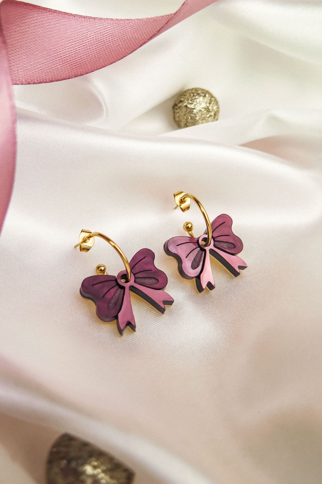 A pair of gold-plated hoop earrings with purple and pink butterfly-shaped charms are displayed on white satin fabric, accented by a pink ribbon and gold decorative balls nearby.