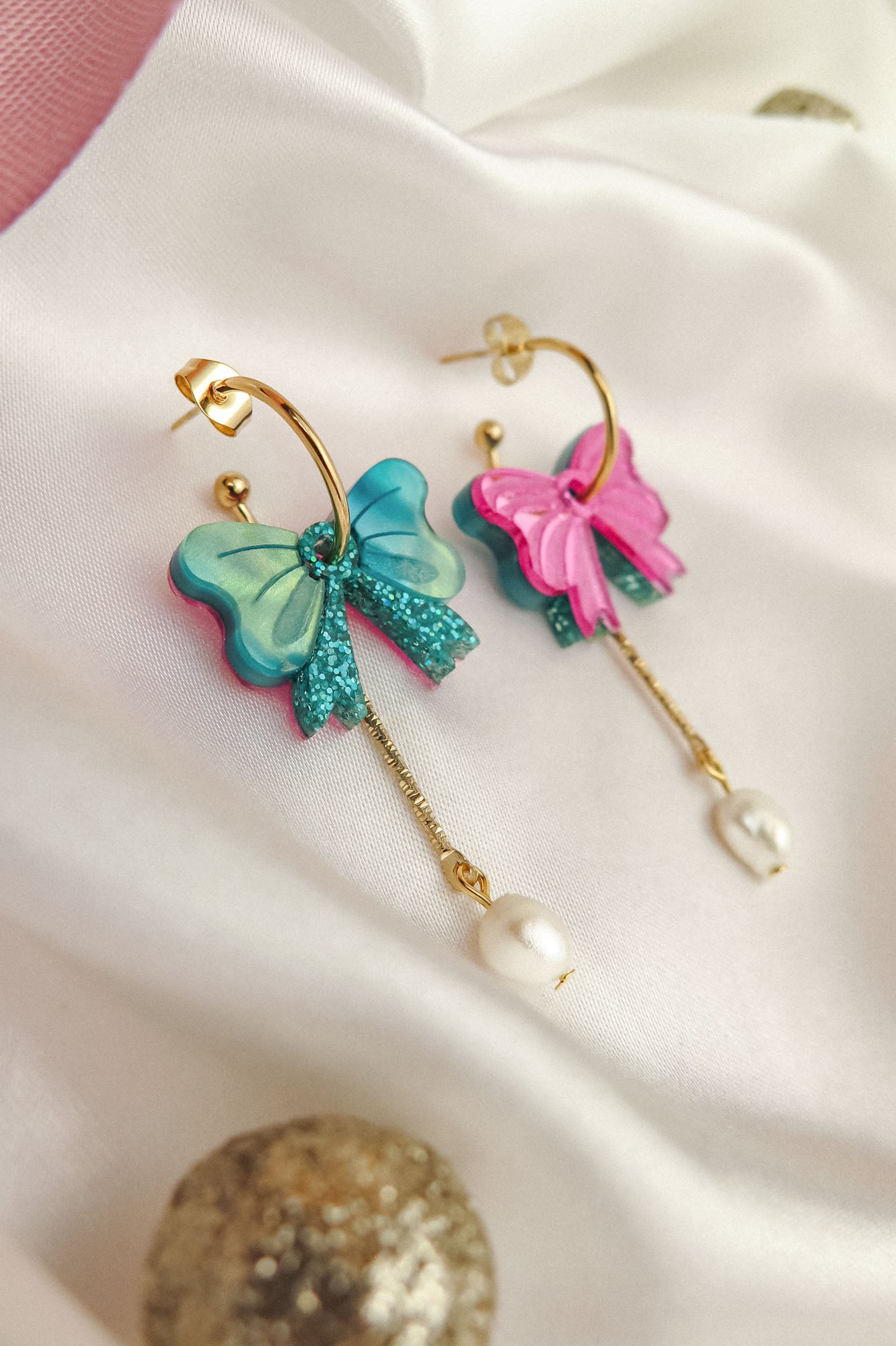 Gold-plated c-hoop earrings with colorful butterfly charms—one blue and one pink—feature freshwater pearl and gold chain accents. These interchangeable dangle earrings are displayed on a white fabric background.