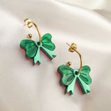 A pair of gold-plated c-hoop earrings with green bow-shaped charms and delicate freshwater pearl accents rests on soft, ivory satin fabric.