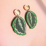 A pair of Calathea Medallion Earrings shaped like green leaves with silver edges and dark green details, set against a soft pink background—perfect for those who love handcrafted statement jewelry inspired by nature.