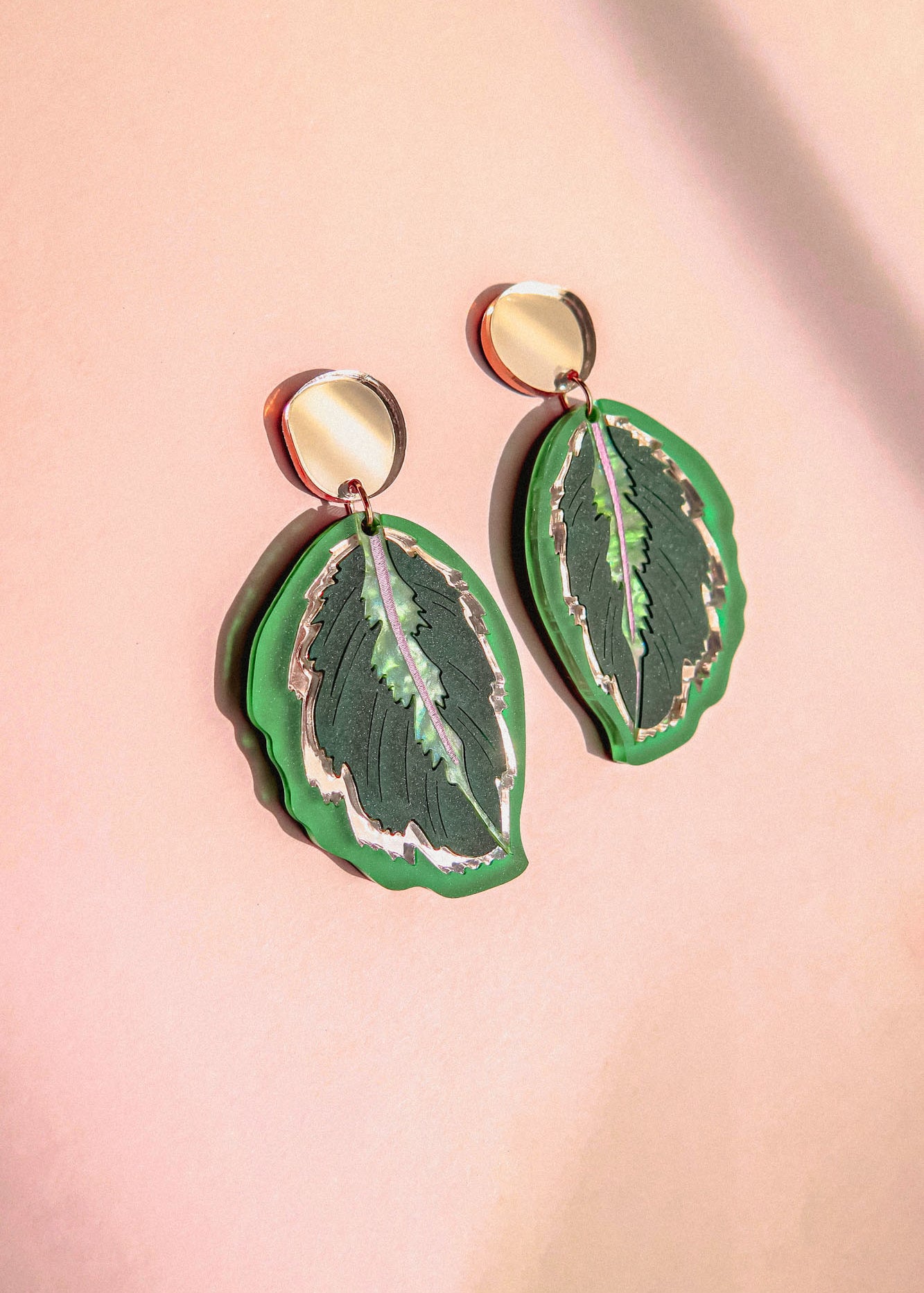 A pair of Calathea Medallion Earrings shaped like green leaves with silver edges and dark green details, set against a soft pink background—perfect for those who love handcrafted statement jewelry inspired by nature.