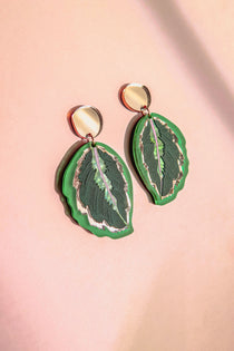 A pair of Calathea Medallion Earrings shaped like green leaves with silver edges and dark green details, set against a soft pink background—perfect for those who love handcrafted statement jewelry inspired by nature.