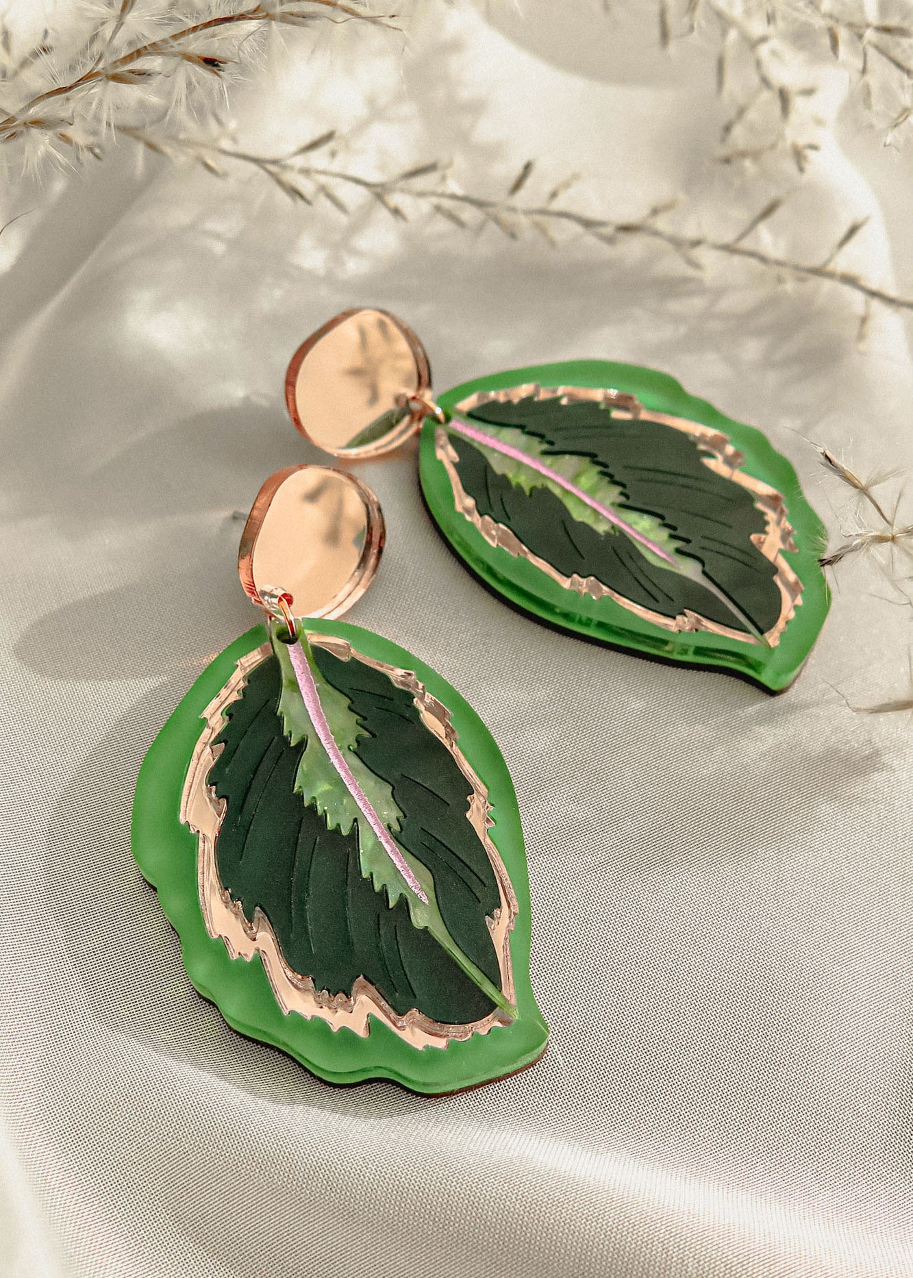 A pair of Calathea Medallion Earrings shaped like green leaves with gold edges and a touch of purple in the center, displayed on a light fabric background with delicate white foliage—nature-inspired jewelry at its finest.