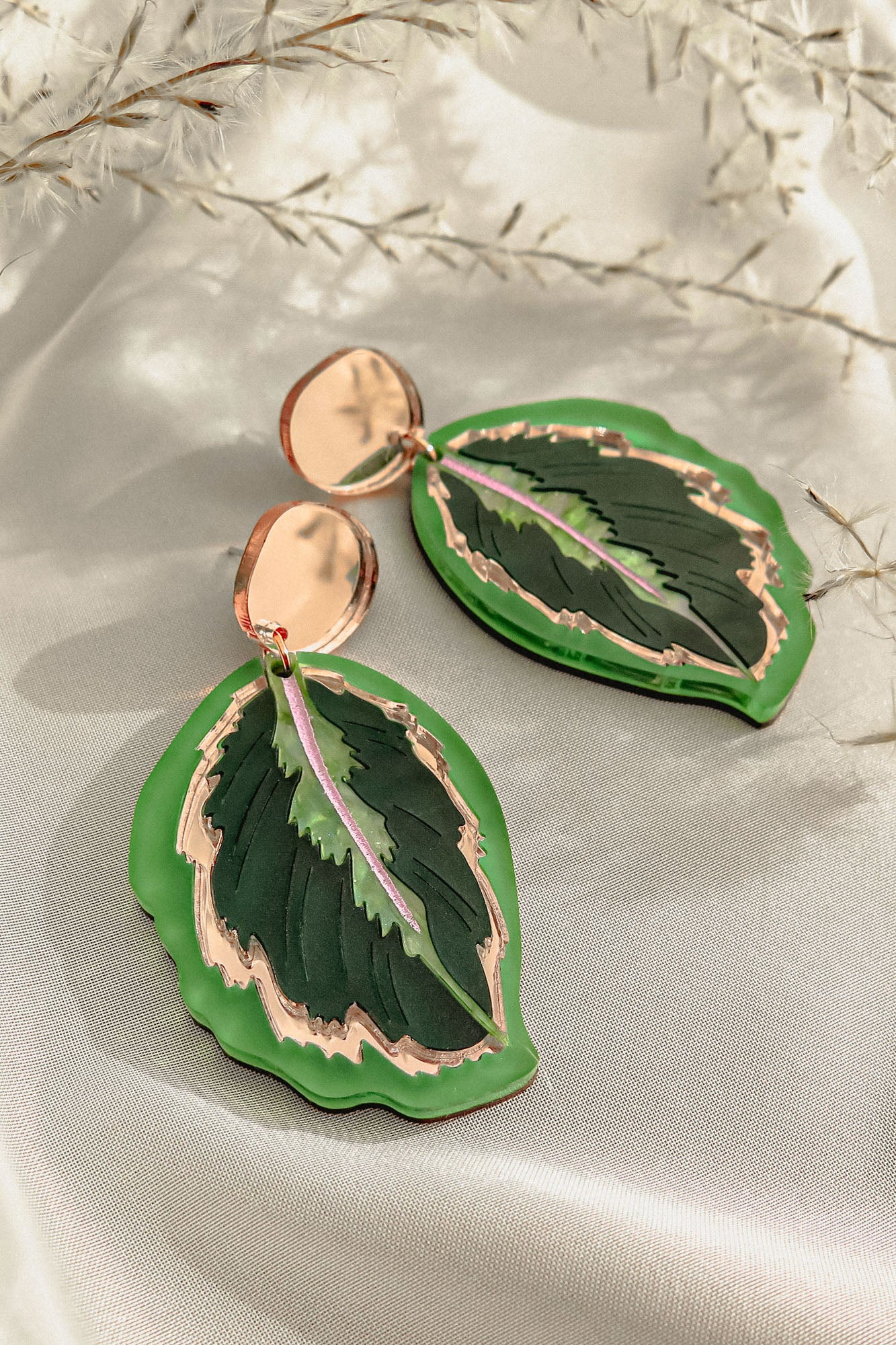 A pair of Calathea Medallion Earrings shaped like green leaves with gold edges and a touch of purple in the center, displayed on a light fabric background with delicate white foliage—nature-inspired jewelry at its finest.