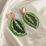 A pair of Calathea Medallion Earrings shaped like green leaves with gold and purple accents, set on a light cream-colored satin fabric background—nature-inspired jewelry perfect for those who love handcrafted statement jewelry.