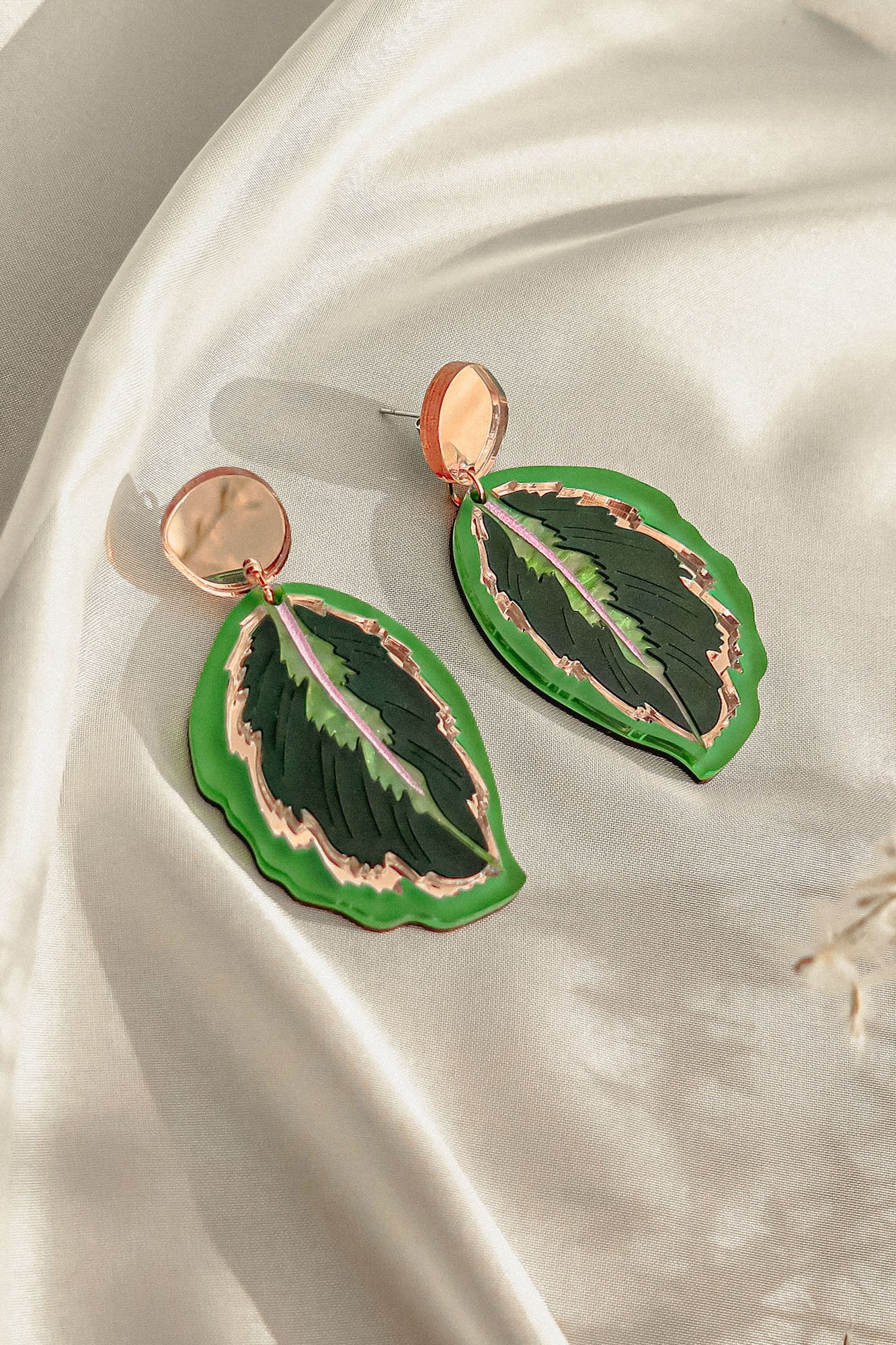 A pair of Calathea Medallion Earrings shaped like green leaves with gold and purple accents, set on a light cream-colored satin fabric background—nature-inspired jewelry perfect for those who love handcrafted statement jewelry.