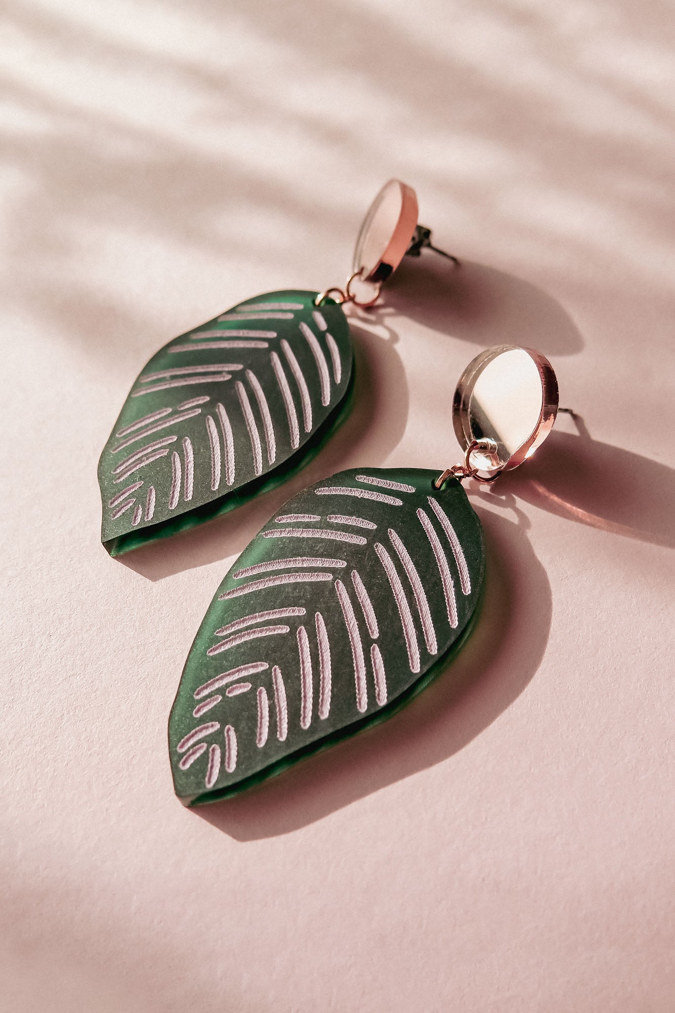 A pair of green, leaf-shaped handcrafted earrings with silver detailing and round metallic studs, inspired by Calathea Ornata Earrings, displayed on a pale pink surface with soft shadows.