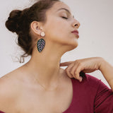A woman with closed eyes and an elegant pose wears a burgundy off-the-shoulder top, a delicate gold necklace, and Calathea Ornata Earrings—handcrafted earrings inspired by tropical nature jewelry. Her dark hair is tied up as she holds a straw hat on her lap.