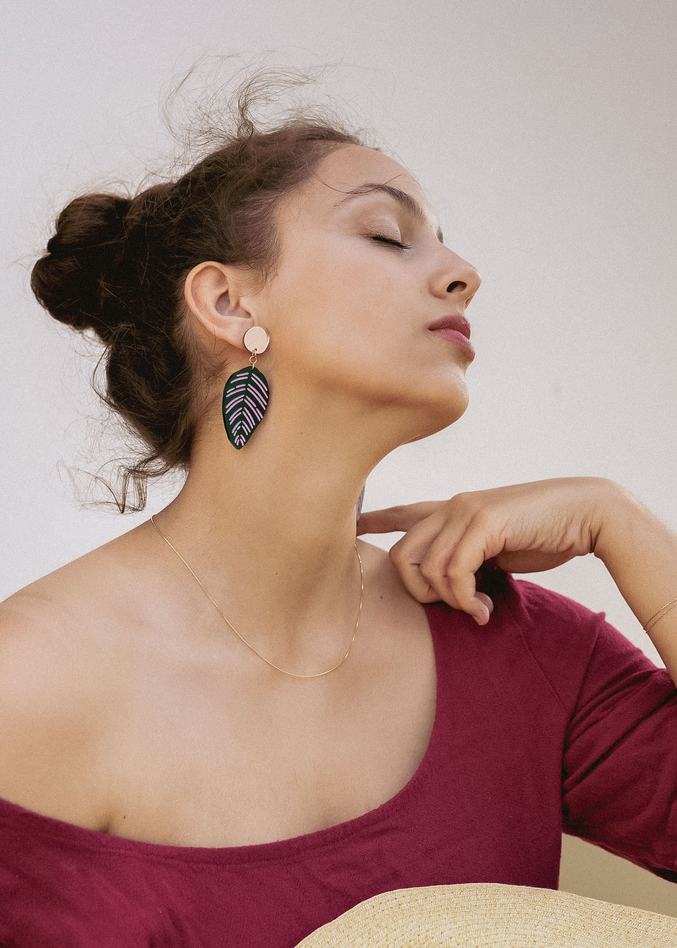 A woman with closed eyes and an elegant pose wears a burgundy off-the-shoulder top, a delicate gold necklace, and Calathea Ornata Earrings—handcrafted earrings inspired by tropical nature jewelry. Her dark hair is tied up as she holds a straw hat on her lap.