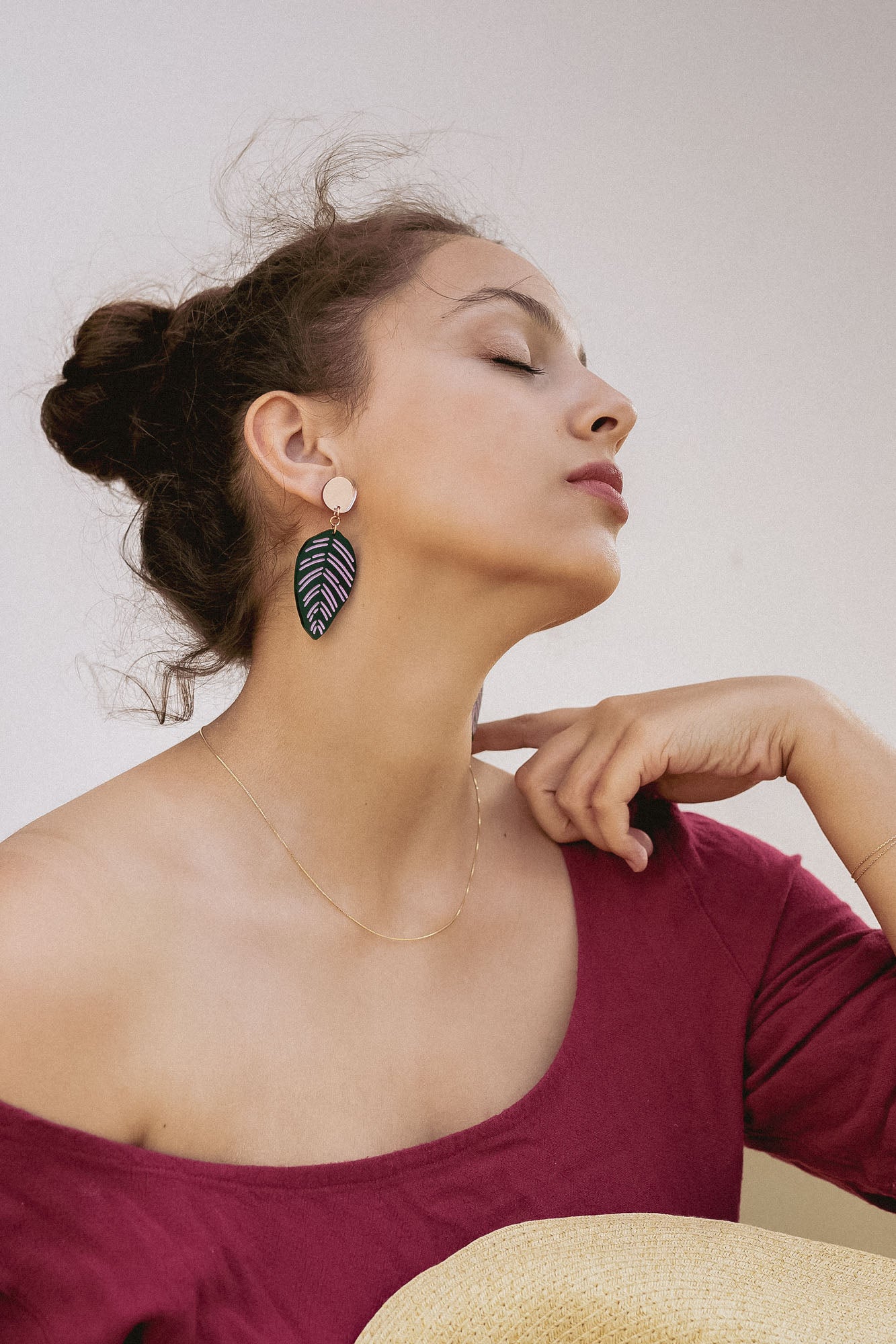 A woman with closed eyes and an elegant pose wears a burgundy off-the-shoulder top, a delicate gold necklace, and Calathea Ornata Earrings—handcrafted earrings inspired by tropical nature jewelry. Her dark hair is tied up as she holds a straw hat on her lap.