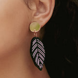 Close-up of a persons ear wearing Calathea Ornata Earrings—handcrafted earrings with a circular gold stud and a black leaf-shaped charm adorned with pink geometric patterns, inspired by tropical nature jewelry. The person has dark hair and wears a maroon top.