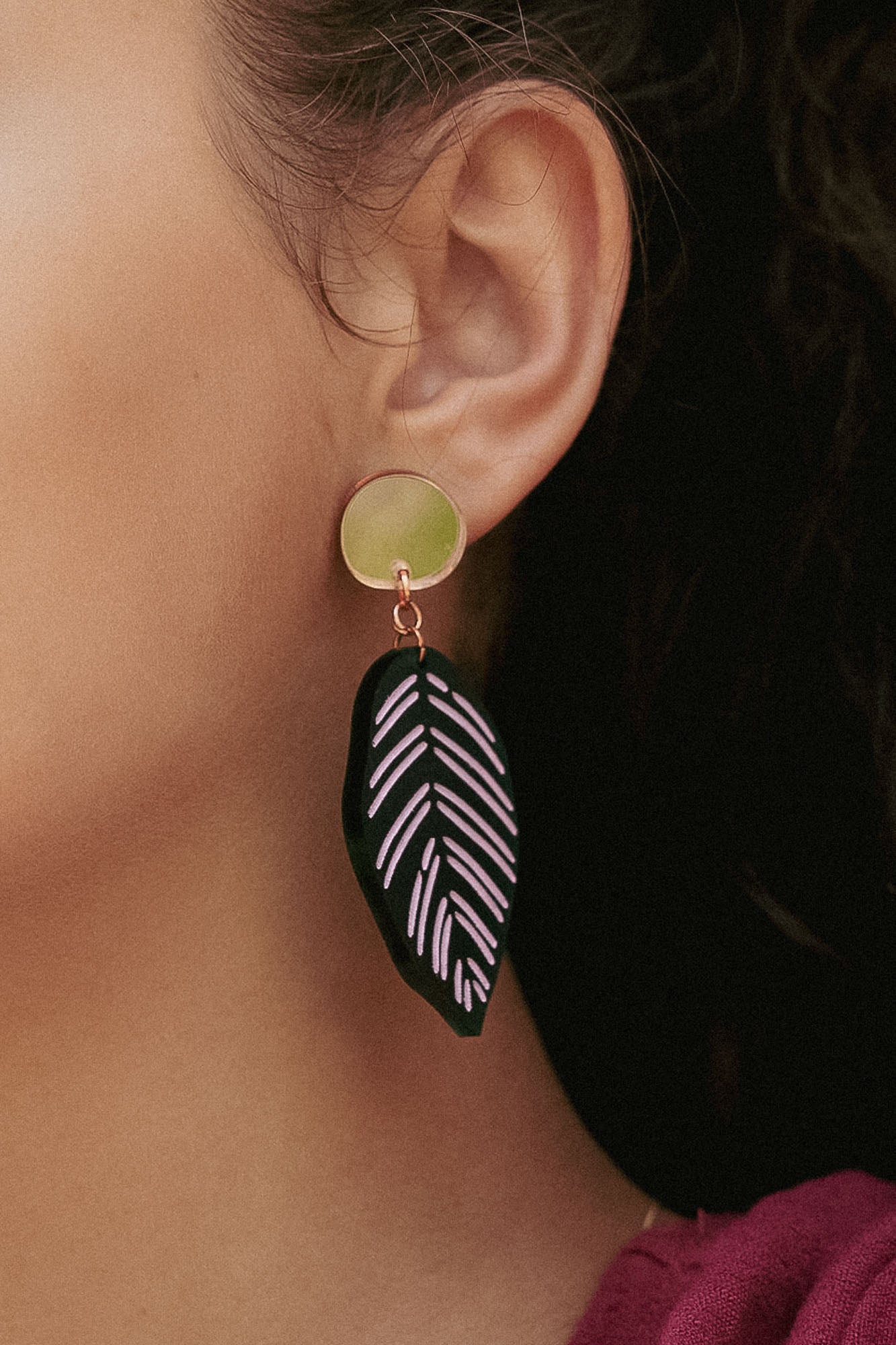 Close-up of a persons ear wearing Calathea Ornata Earrings—handcrafted earrings with a circular gold stud and a black leaf-shaped charm adorned with pink geometric patterns, inspired by tropical nature jewelry. The person has dark hair and wears a maroon top.