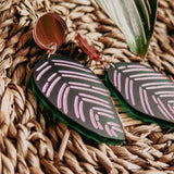 A pair of leaf-shaped, handcrafted earrings with pink and white line designs rest on a woven straw surface beside a green leaf with light stripes, echoing the beauty of Calathea Ornata Earrings and tropical nature jewelry.