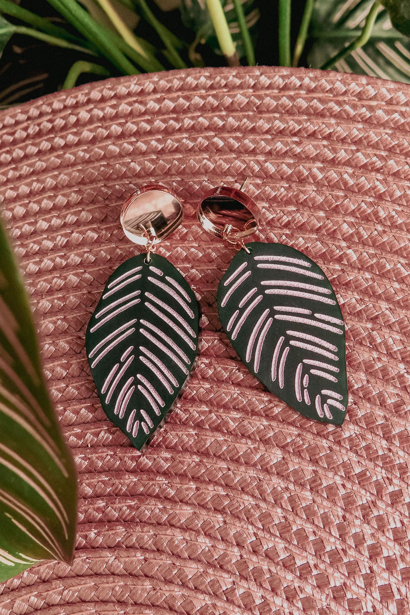 A pair of handcrafted earrings, the Calathea Ornata Earrings, with metallic round studs and dark green leaf-shaped pendants featuring light pink vein patterns, are displayed on a textured pink woven surface with green leaves in the background—a true piece of tropical nature jewelry.