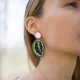 Close-up of a woman wearing Calathea Medallion Earrings—handcrafted statement jewelry in green and black with a gold edge. She has light brown hair, a mauve top, and only the right side of her face is visible.