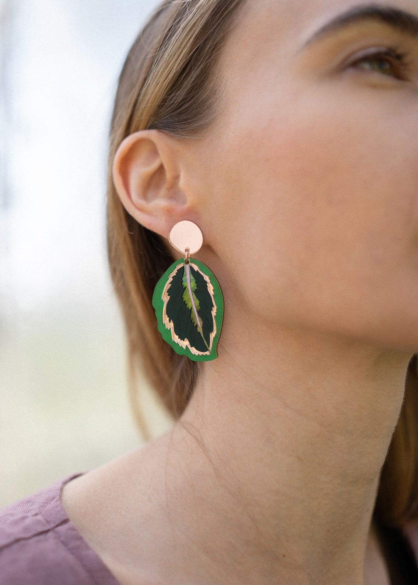 Close-up of a woman wearing Calathea Medallion Earrings—handcrafted statement jewelry in green and black with a gold edge. She has light brown hair, a mauve top, and only the right side of her face is visible.