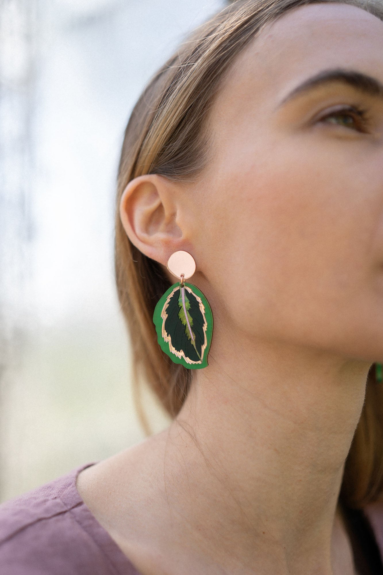 Close-up of a woman wearing Calathea Medallion Earrings—handcrafted statement jewelry in green and black with a gold edge. She has light brown hair, a mauve top, and only the right side of her face is visible.