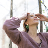 A woman in a mauve buttoned top stands outdoors by a glass wall, eyes closed and hands touching her forehead, wearing Calathea Medallion Earrings—nature-inspired jewelry that brings a relaxed elegance to her look in natural light.