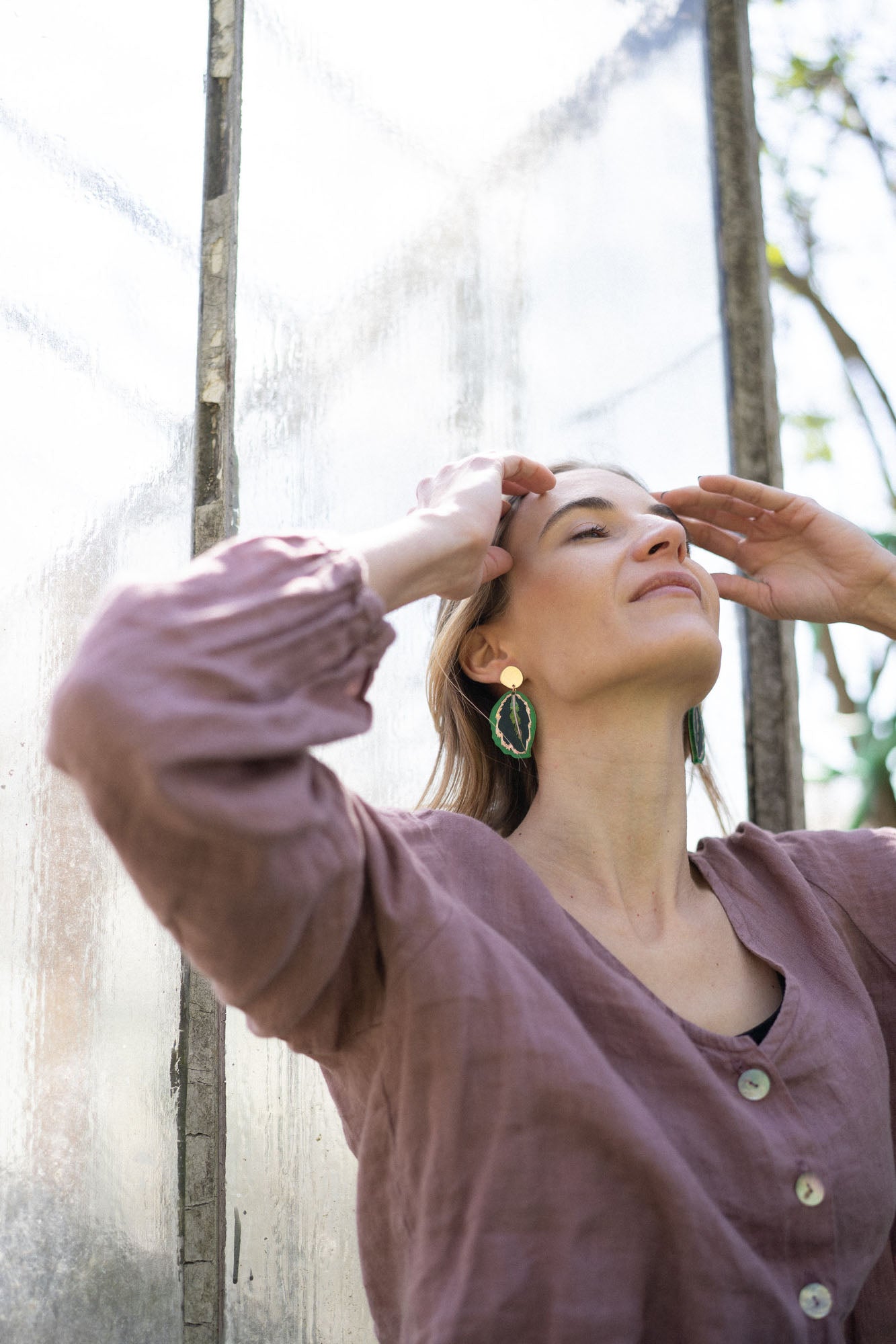 A woman in a mauve buttoned top stands outdoors by a glass wall, eyes closed and hands touching her forehead, wearing Calathea Medallion Earrings—nature-inspired jewelry that brings a relaxed elegance to her look in natural light.