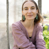 A woman with light brown hair, wearing a mauve blouse and Calathea Medallion Earrings, sits outdoors in sunlight, smiling softly at the camera. Blurred greenery and a glass panel create a serene backdrop for her nature-inspired jewelry.