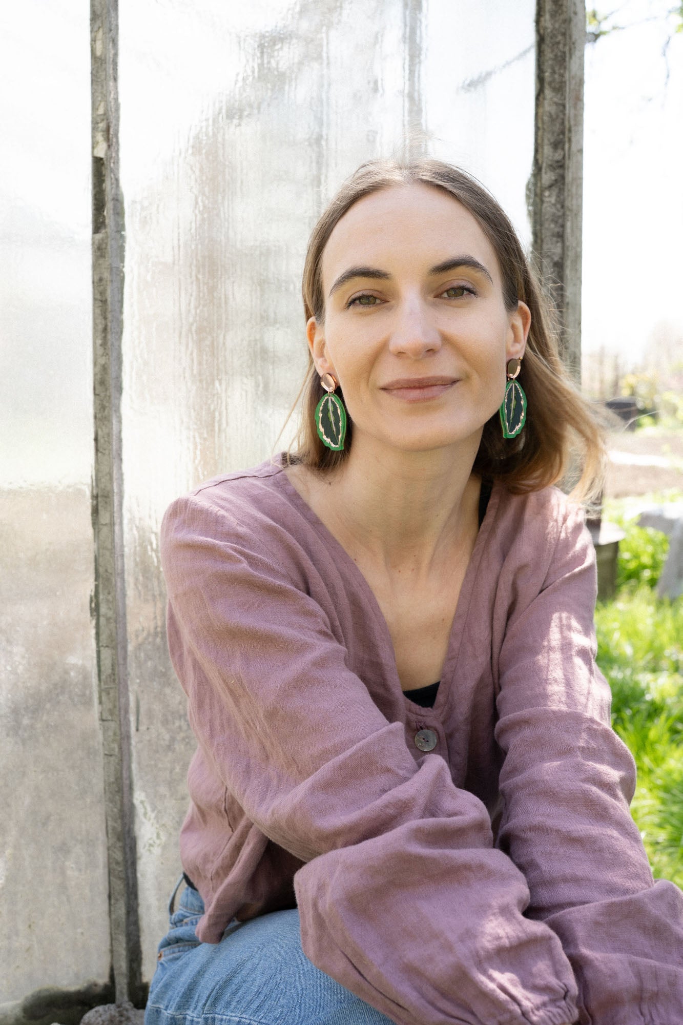 A woman with light brown hair, wearing a mauve blouse and Calathea Medallion Earrings, sits outdoors in sunlight, smiling softly at the camera. Blurred greenery and a glass panel create a serene backdrop for her nature-inspired jewelry.