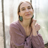 A woman with light brown hair, wearing a mauve blouse and green Calathea Medallion Earrings, sits by a sunlit window, smiling gently at the camera, her nature-inspired jewelry adding a unique touch to her look.