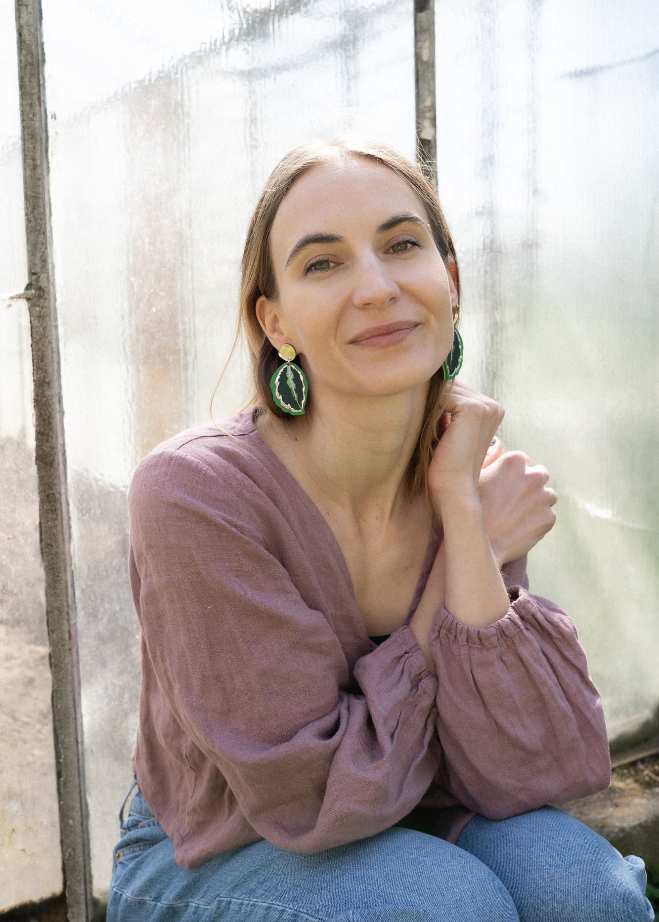 A woman with light brown hair, wearing a mauve blouse and green Calathea Medallion Earrings, sits by a sunlit window, smiling gently at the camera, her nature-inspired jewelry adding a unique touch to her look.