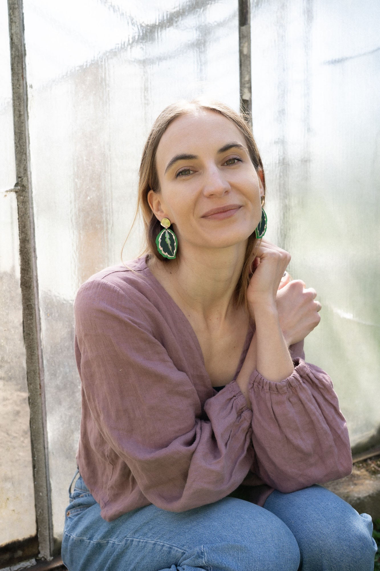 A woman with light brown hair, wearing a mauve blouse and green Calathea Medallion Earrings, sits by a sunlit window, smiling gently at the camera, her nature-inspired jewelry adding a unique touch to her look.