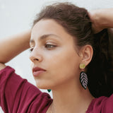 A woman with wavy brown hair pulled back wears a burgundy top, a delicate gold necklace, and Calathea Ornata Earrings—handcrafted earrings inspired by tropical nature jewelry—featuring green beads and a black-and-white leaf design.