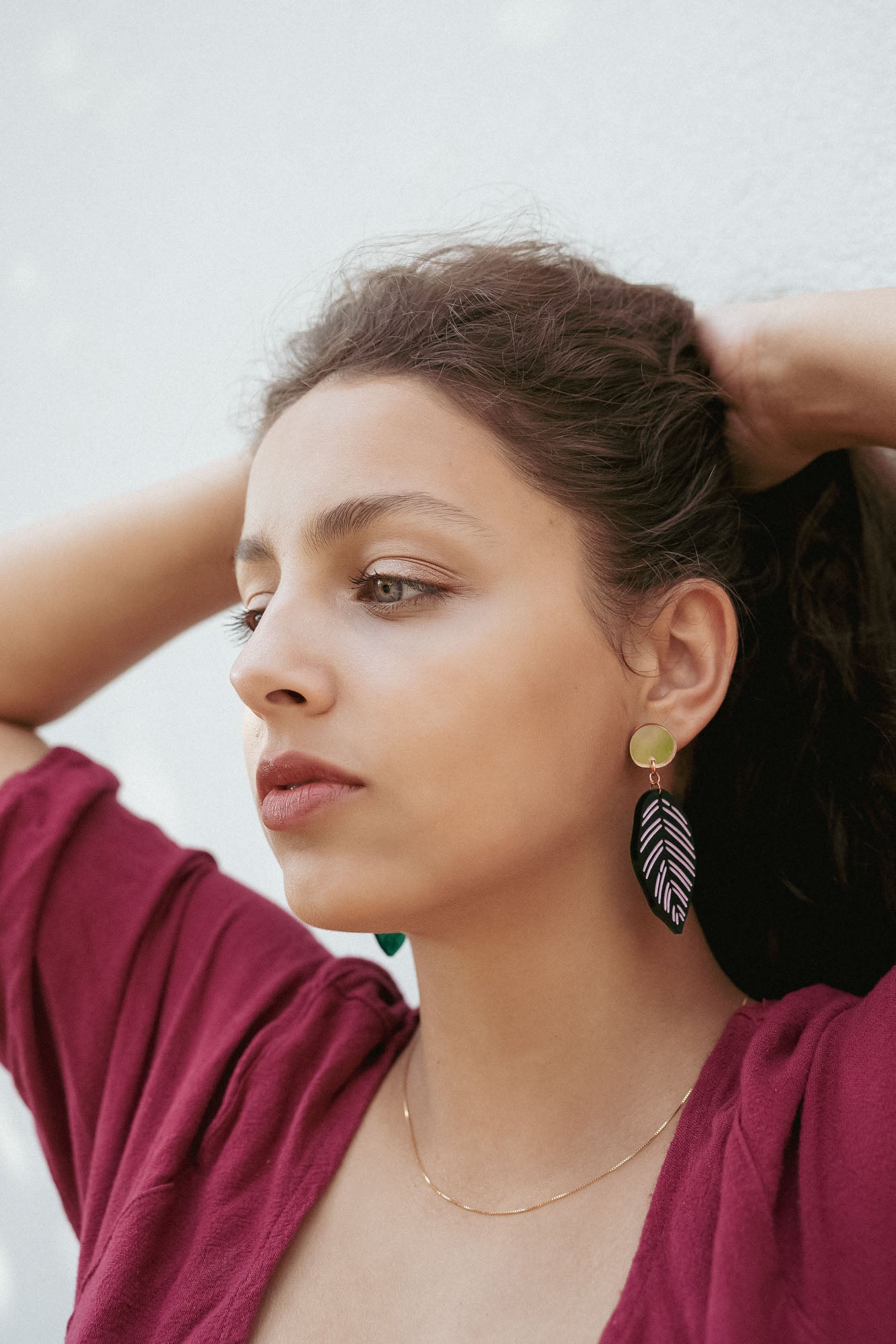 A woman with wavy brown hair pulled back wears a burgundy top, a delicate gold necklace, and Calathea Ornata Earrings—handcrafted earrings inspired by tropical nature jewelry—featuring green beads and a black-and-white leaf design.