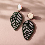 A pair of handcrafted earrings with round gold studs and green leaf-shaped pendants with white vein details, inspired by Calathea Ornata Earrings, displayed on a pink surface with soft shadows.