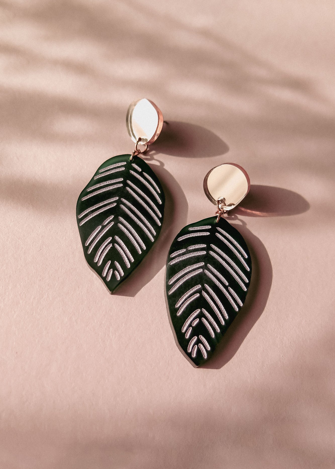 A pair of handcrafted earrings with round gold studs and green leaf-shaped pendants with white vein details, inspired by Calathea Ornata Earrings, displayed on a pink surface with soft shadows.