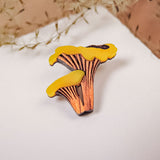 A yellow and brown handcrafted Chanterelle Mushroom Pin sits on a white surface, with soft dried grass in the background.