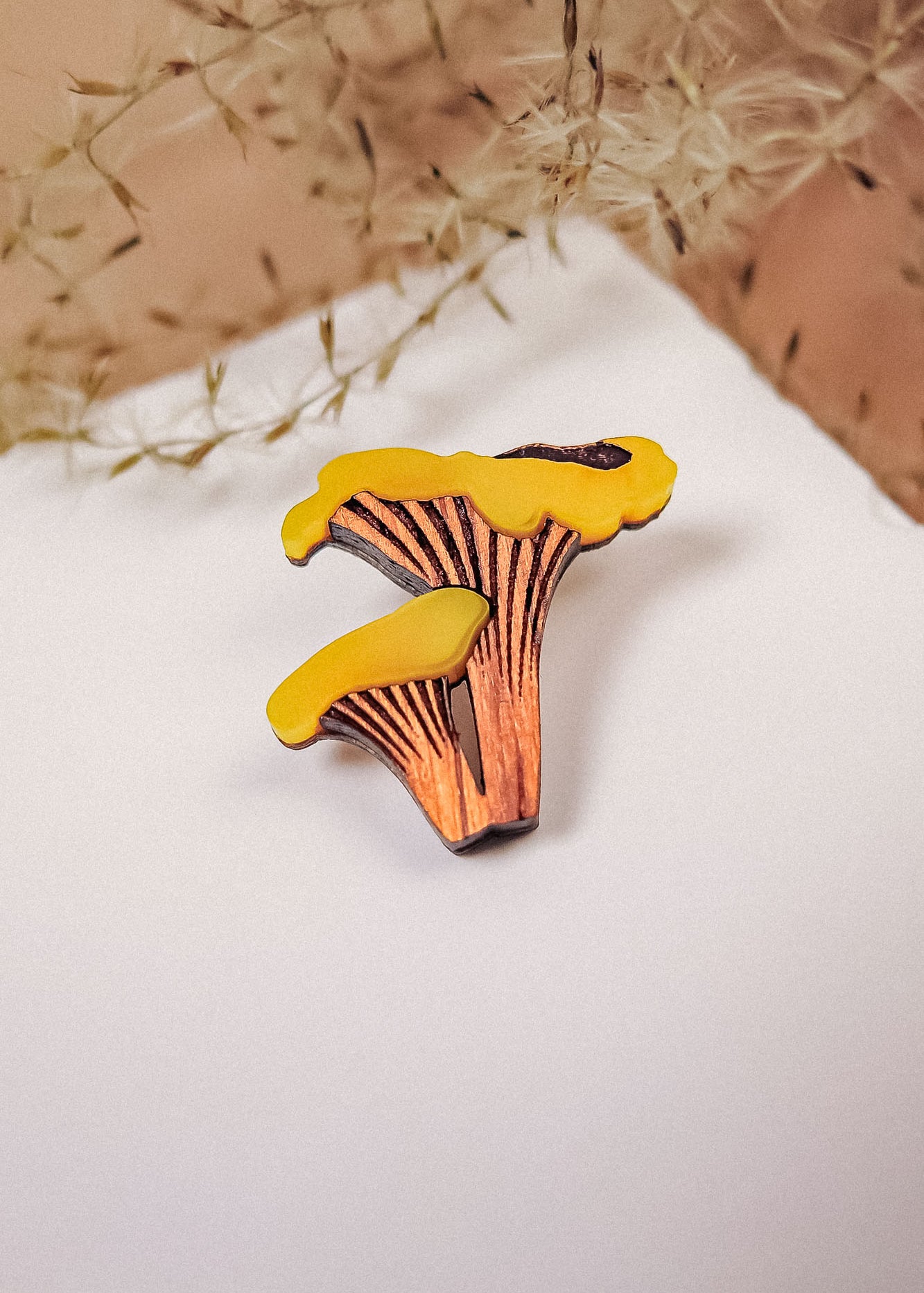 A yellow and brown handcrafted Chanterelle Mushroom Pin sits on a white surface, with soft dried grass in the background.