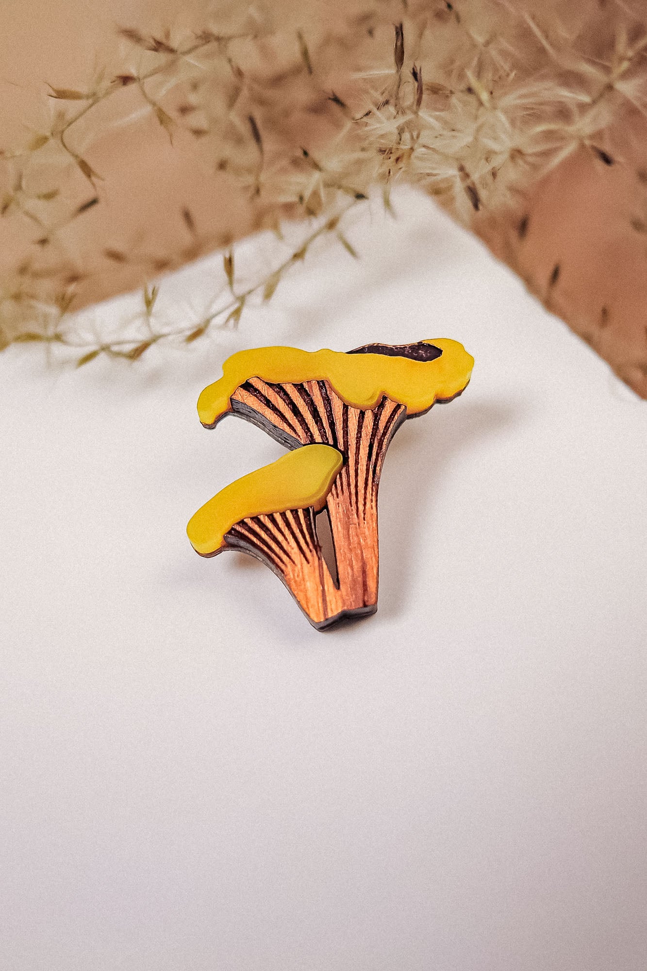 A yellow and brown handcrafted Chanterelle Mushroom Pin sits on a white surface, with soft dried grass in the background.